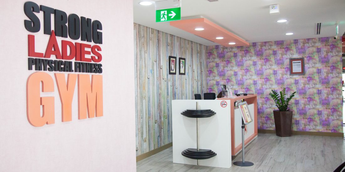 The modern and brightly lit reception area of a 'STRONG LADIES PHYSICAL FITNESS GYM', featuring a unique desk with weights, textured walls, and a welcoming ambiance, designed by commercial fit-out specialists in UAE.