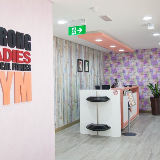 The modern and brightly lit reception area of a 'STRONG LADIES PHYSICAL FITNESS GYM', featuring a unique desk with weights, textured walls, and a welcoming ambiance, designed by commercial fit-out specialists in UAE.