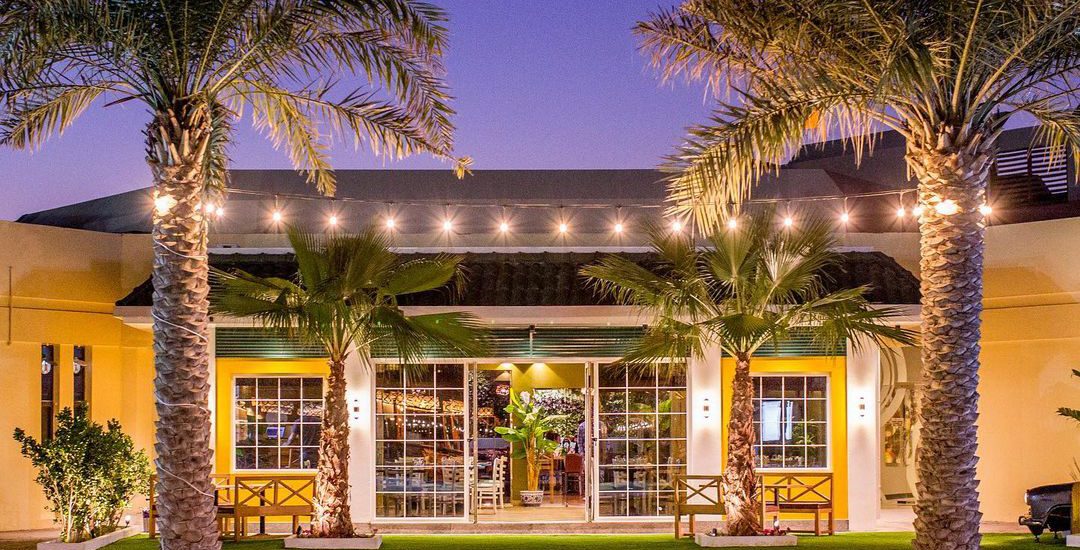 A warmly lit commercial building at twilight, with large windows, string lights, and tall palm trees, seen across a swimming pool, highlighting Seven Star's commercial fit-out specialists in UAE expertise in creating inviting exteriors.