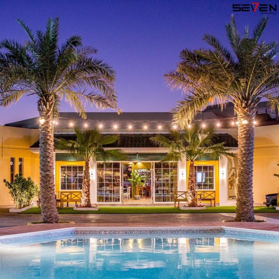 A warmly lit commercial building at twilight, with large windows, string lights, and tall palm trees, seen across a swimming pool, highlighting Seven Star's commercial fit-out specialists in UAE expertise in creating inviting exteriors.