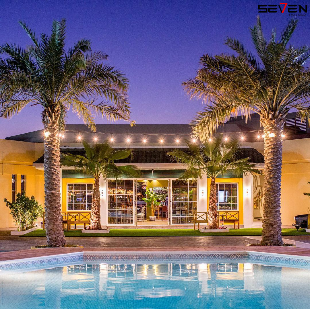 A warmly lit commercial building at twilight, with large windows, string lights, and tall palm trees, seen across a swimming pool, highlighting Seven Star's commercial fit-out specialists in UAE expertise in creating inviting exteriors.