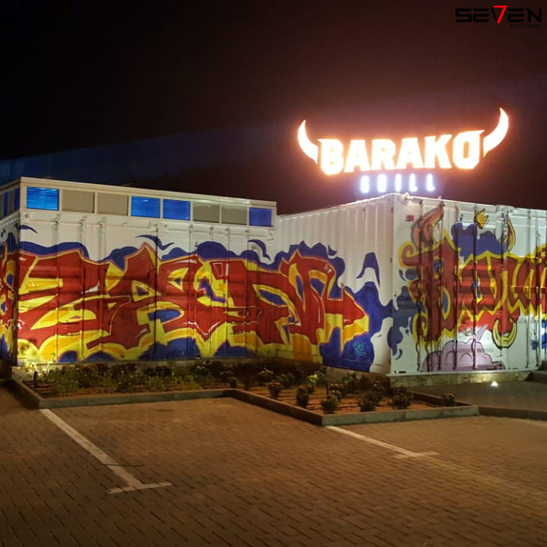 The vibrant exterior of Barako Grill at night, designed by Seven Star in Dubai, featuring shipping containers covered in colorful graffiti and an illuminated bull-horn sign, showcasing their expertise as commercial fit-out specialists in UAE.